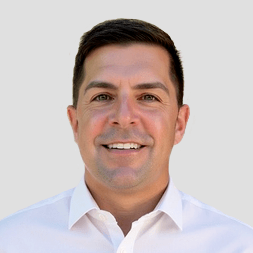 A man with short dark hair and a light complexion is smiling. He is wearing a white collared shirt and is pictured against a plain, light gray background.