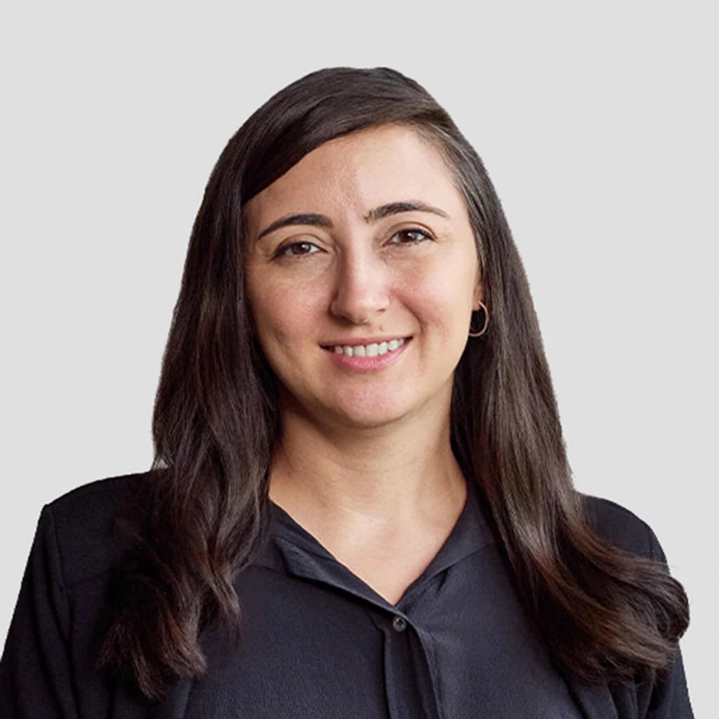 A woman with long, wavy dark hair, wearing a black blouse and small hoop earrings, smiles at the camera against a plain light gray background.