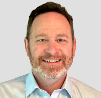 Smiling bearded man with short brown hair wearing a light blue collared shirt, posing in front of a plain light gray background.