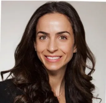 A woman with long, wavy brown hair smiles at the camera. She is wearing a black blazer and is posed against a plain, light background.