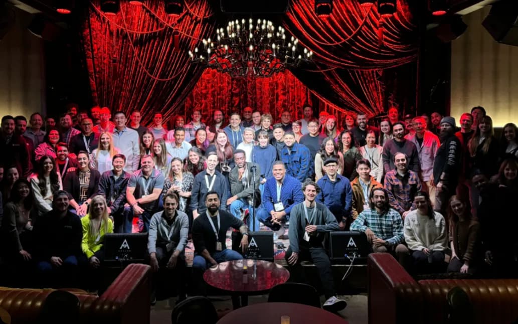 A large group of people pose together on a stage in front of red curtains and a chandelier, with some seated in velvet chairs and others standing behind them, smiling for a group photo in a dimly lit venue.