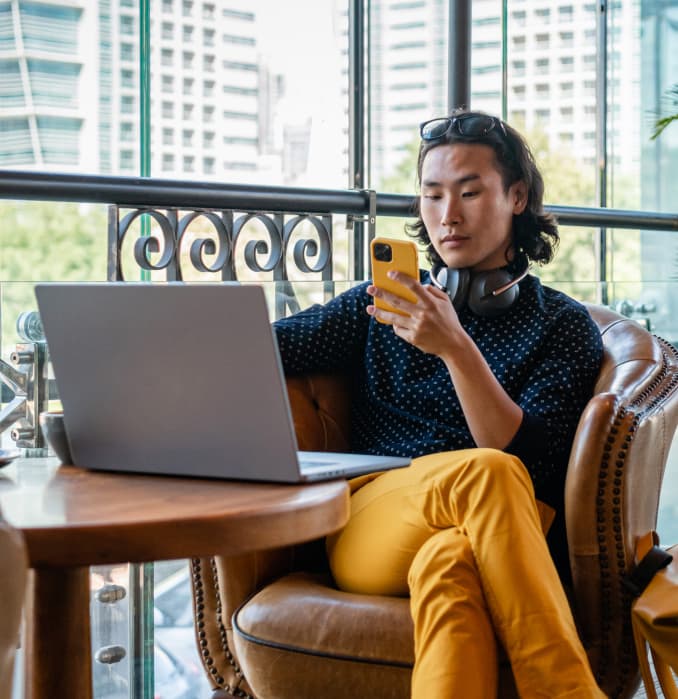 A person sits in a café on a leather chair, holding a yellow phone and looking at it, with ai agents displayed on their laptop open on the wooden table. Headphones rest around their neck, city buildings visible through large windows.