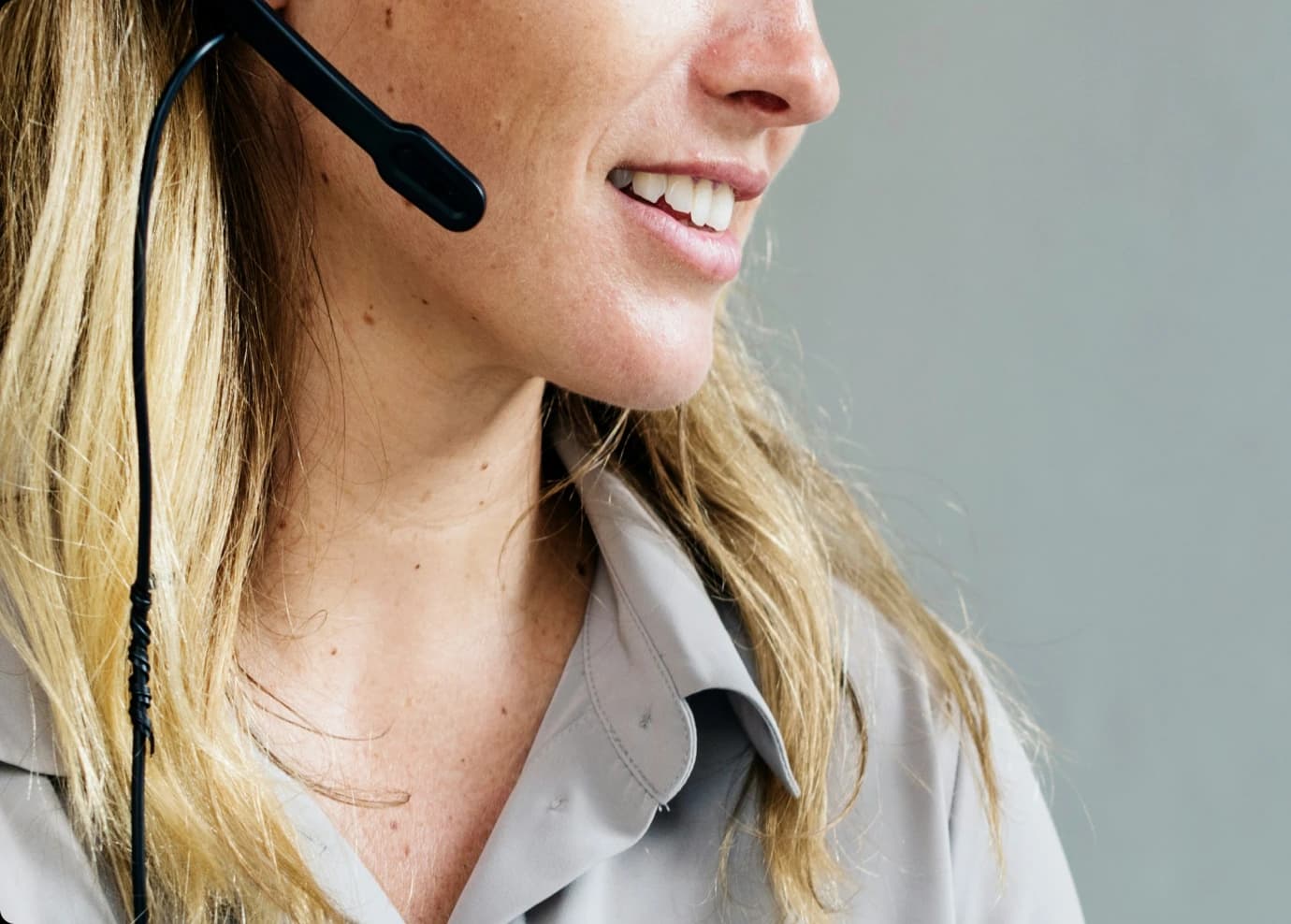 Close-up of a woman with blonde hair smiling while wearing a headset and a light gray collared shirt, suggesting she is working in customer service or support.