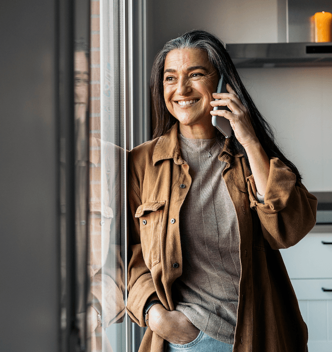 A smiling woman with long gray hair stands by a window, talking on her smartphone. She wears a brown jacket over a beige top and has one hand in her pocket in a modern kitchen.