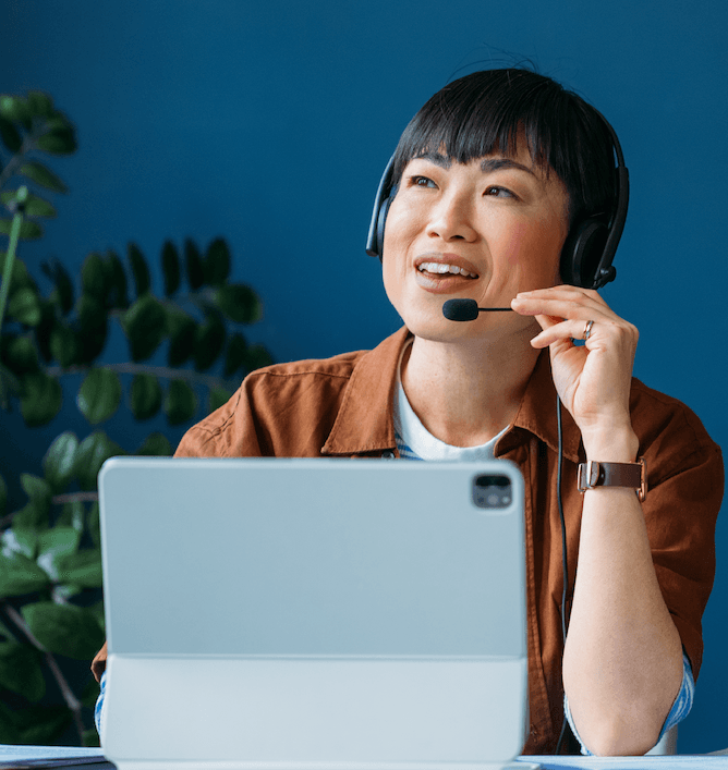 A person wearing a headset sits at a desk, smiling and looking up while speaking. They are in front of an open laptop, with a leafy plant visible in the background against a blue wall.