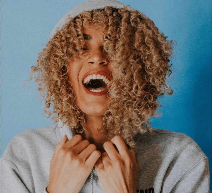 A person with curly hair smiles widely, pulling up the hood of a light gray sweatshirt against a blue background.