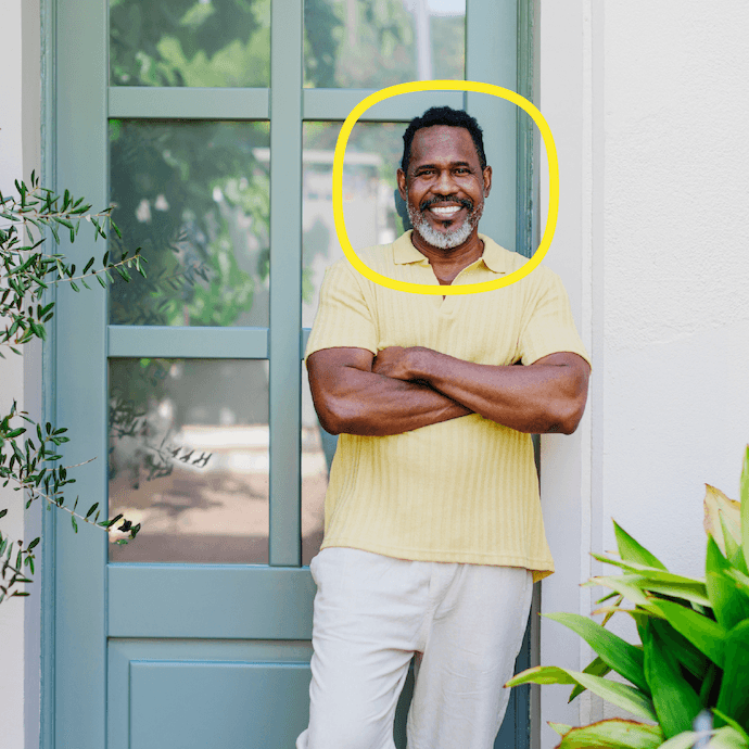 A man in a yellow shirt and light pants stands smiling with arms crossed in front of a blue door, surrounded by green plants. His face is highlighted by a yellow oval.