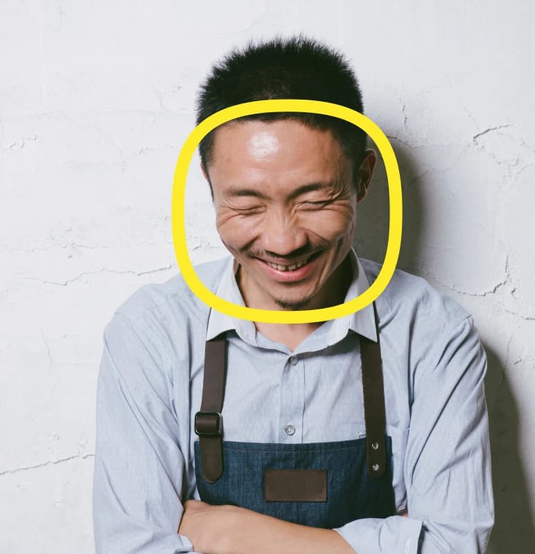 A man wearing a blue apron and light shirt stands against a white wall, smiling with his eyes closed. A yellow circle is drawn around his face.