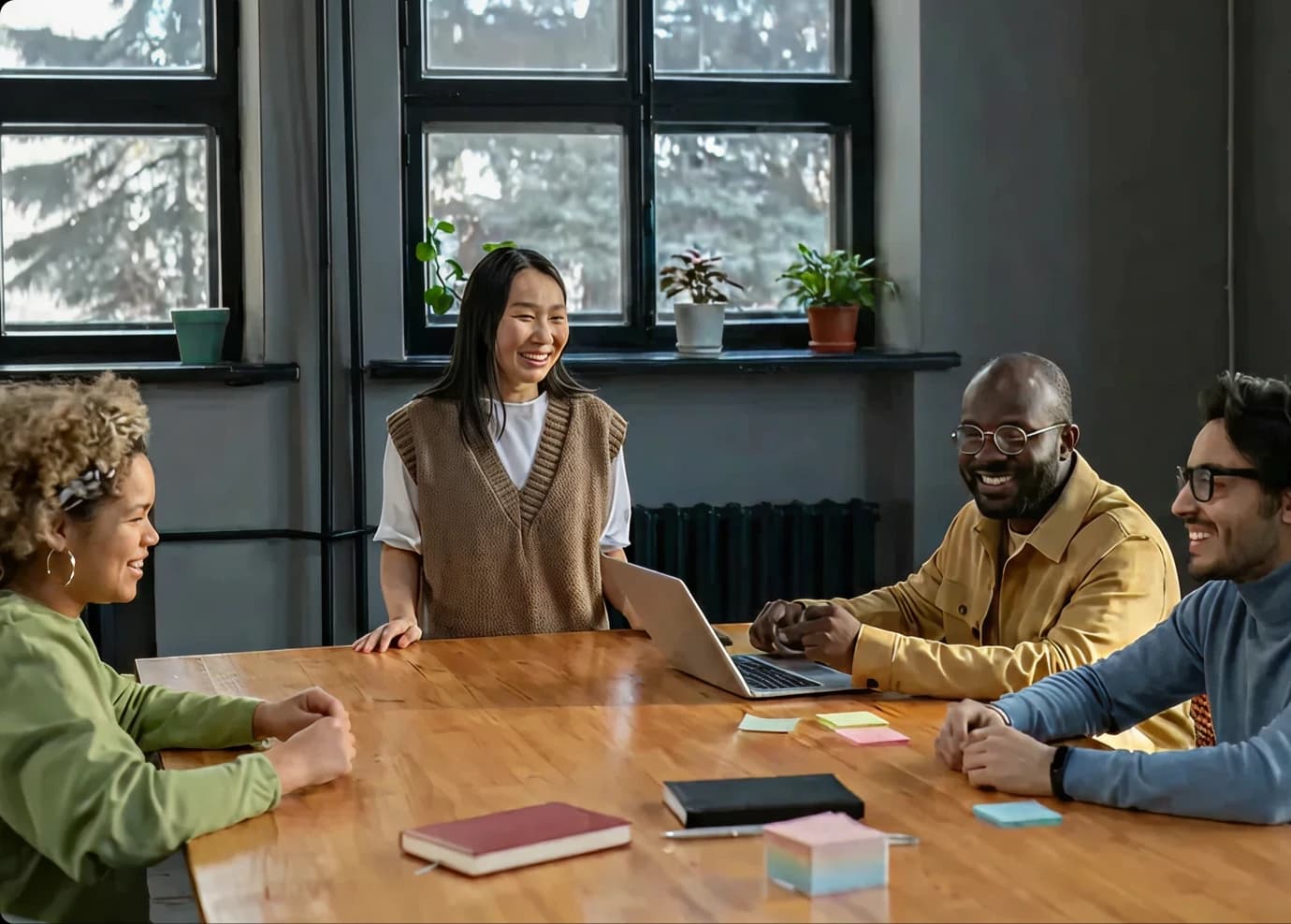 Four people sit at a wooden table with notebooks, sticky notes, and a laptop, while a woman stands smiling. They are in a bright room with plants and large windows. Everyone appears engaged and happy.