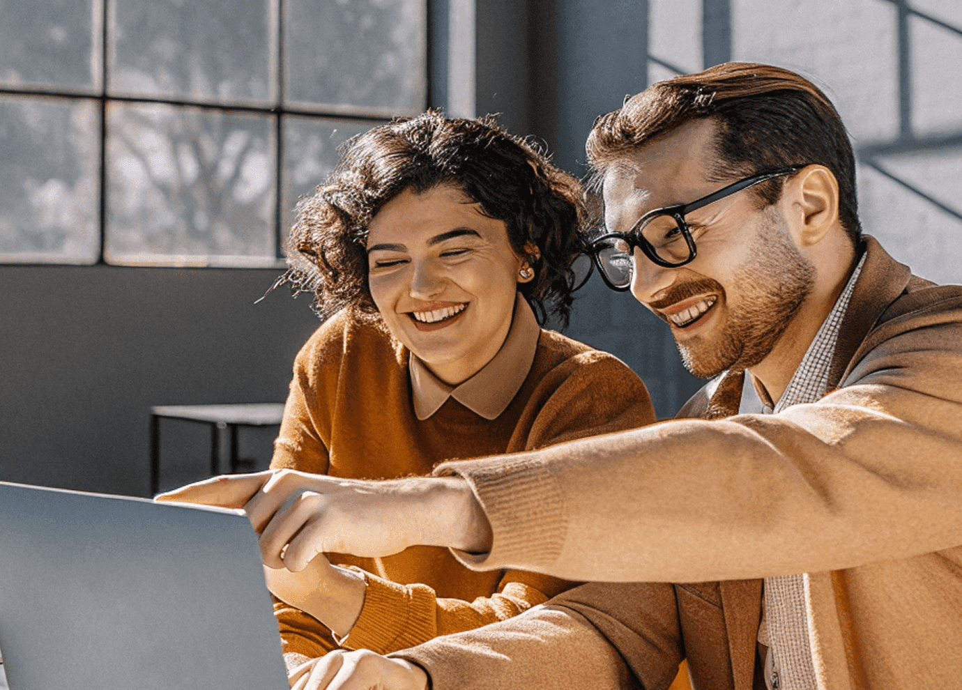 Two people sit together at a desk, smiling and looking at a laptop screen. One is pointing at the screen, and they appear to be enjoying a collaborative discussion in a sunlit, modern office space.