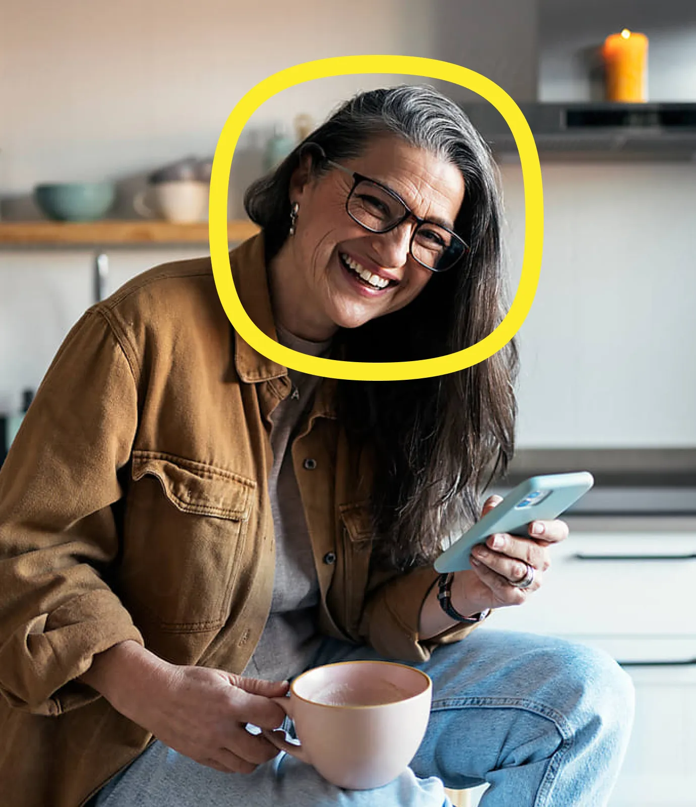 A smiling woman with long gray hair and glasses holds a phone and a mug in a cozy kitchen. A yellow circle is drawn around her head, highlighting her face.