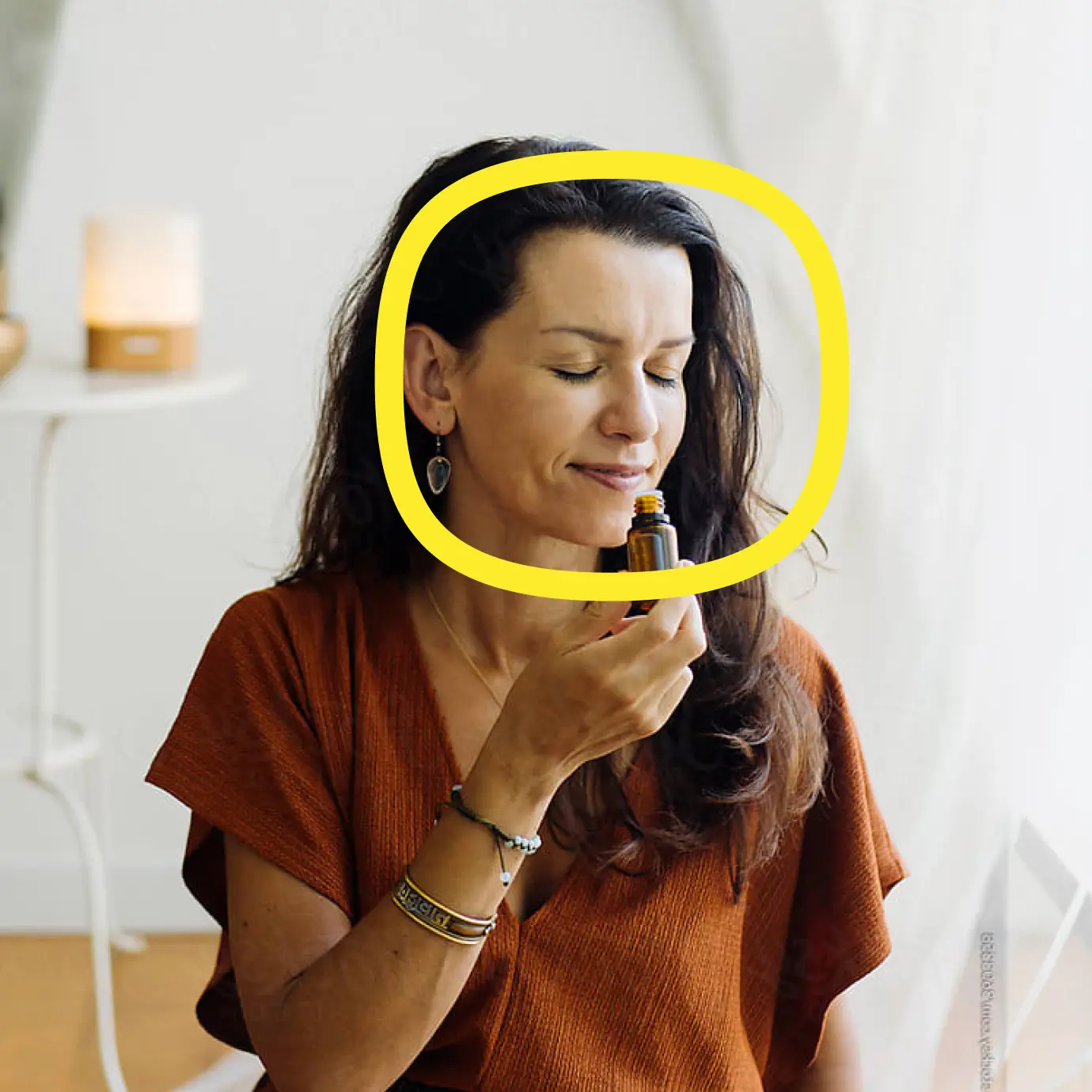 A woman with long dark hair, wearing a rust-colored top, sits with her eyes closed, holding a small dropper bottle near her face. A yellow circle highlights her face. The background is softly lit and minimal.