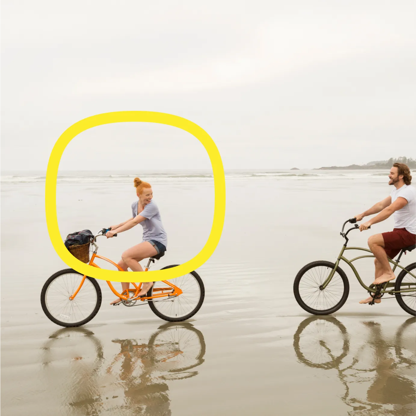Two people ride bicycles on a wet, sandy beach. The person on the left, riding an orange bike, is highlighted by a yellow outline. The sky is overcast and the ocean is in the background.