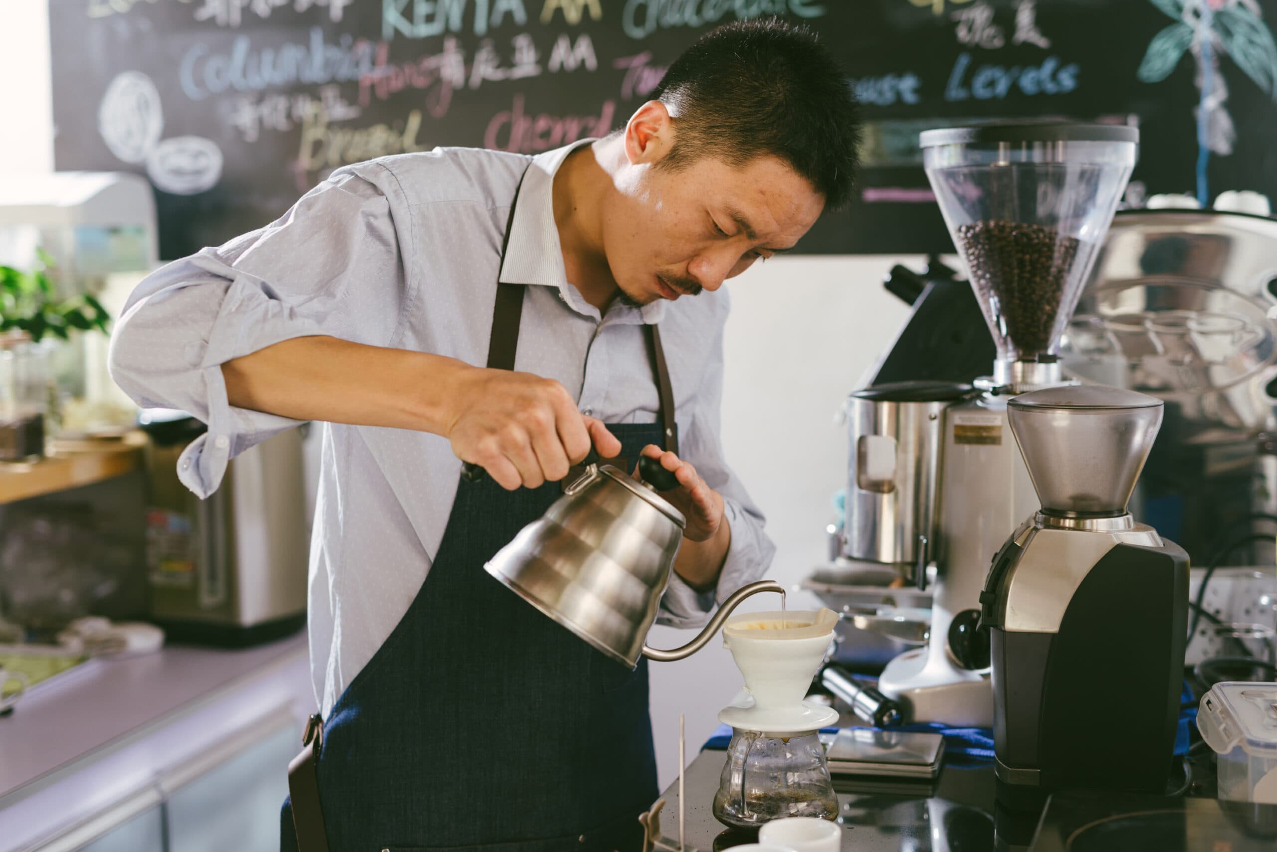A barista wearing an apron carefully pours hot water from a kettle over a coffee filter in a café.