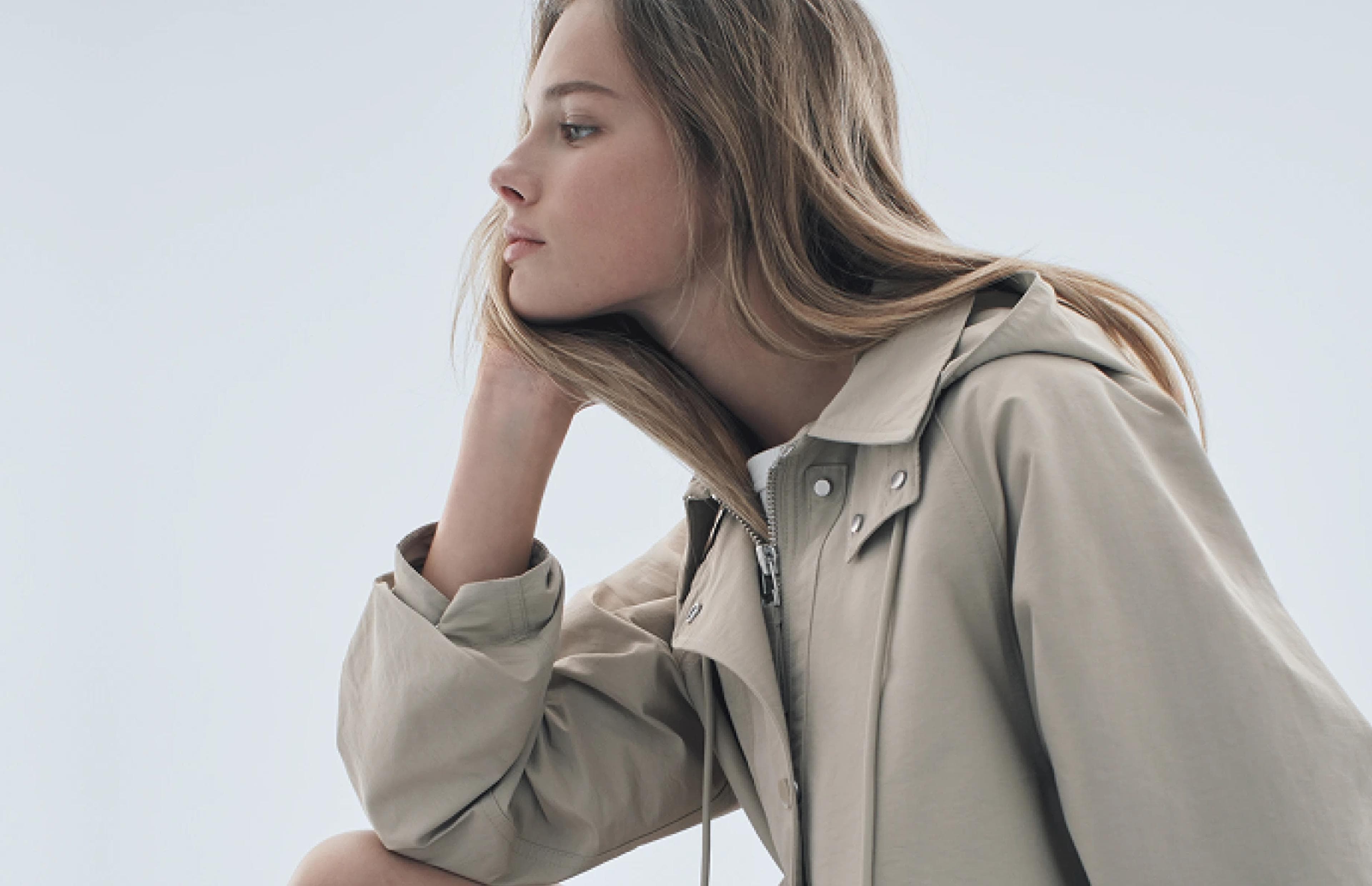 A young woman with light brown hair looks thoughtfully into the distance, resting her chin on her hand. She is wearing a beige trench coat over a light shirt against a pale, neutral background.