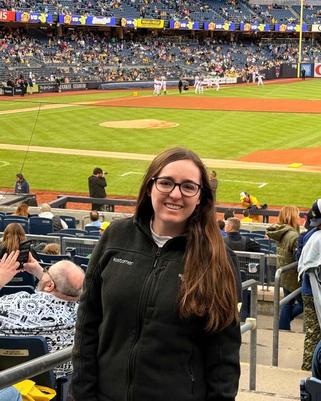 A woman with long brown hair and glasses smiles at the camera in a stadium seat at a baseball game, capturing authentic fan experiences with the field, players, and an enthusiastic crowd in the background.
