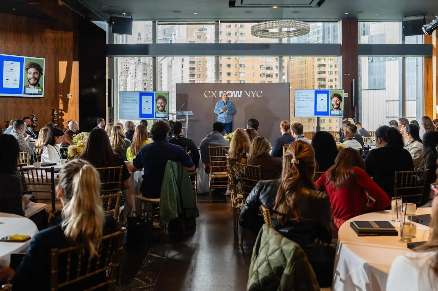 Photo of an event called CX Now NYC with Kustomer CEO Brad Birnbaum on stage with three screens and attendees sitting at round tables watching, against a backdrop of floor to ceiling windows and a city view.