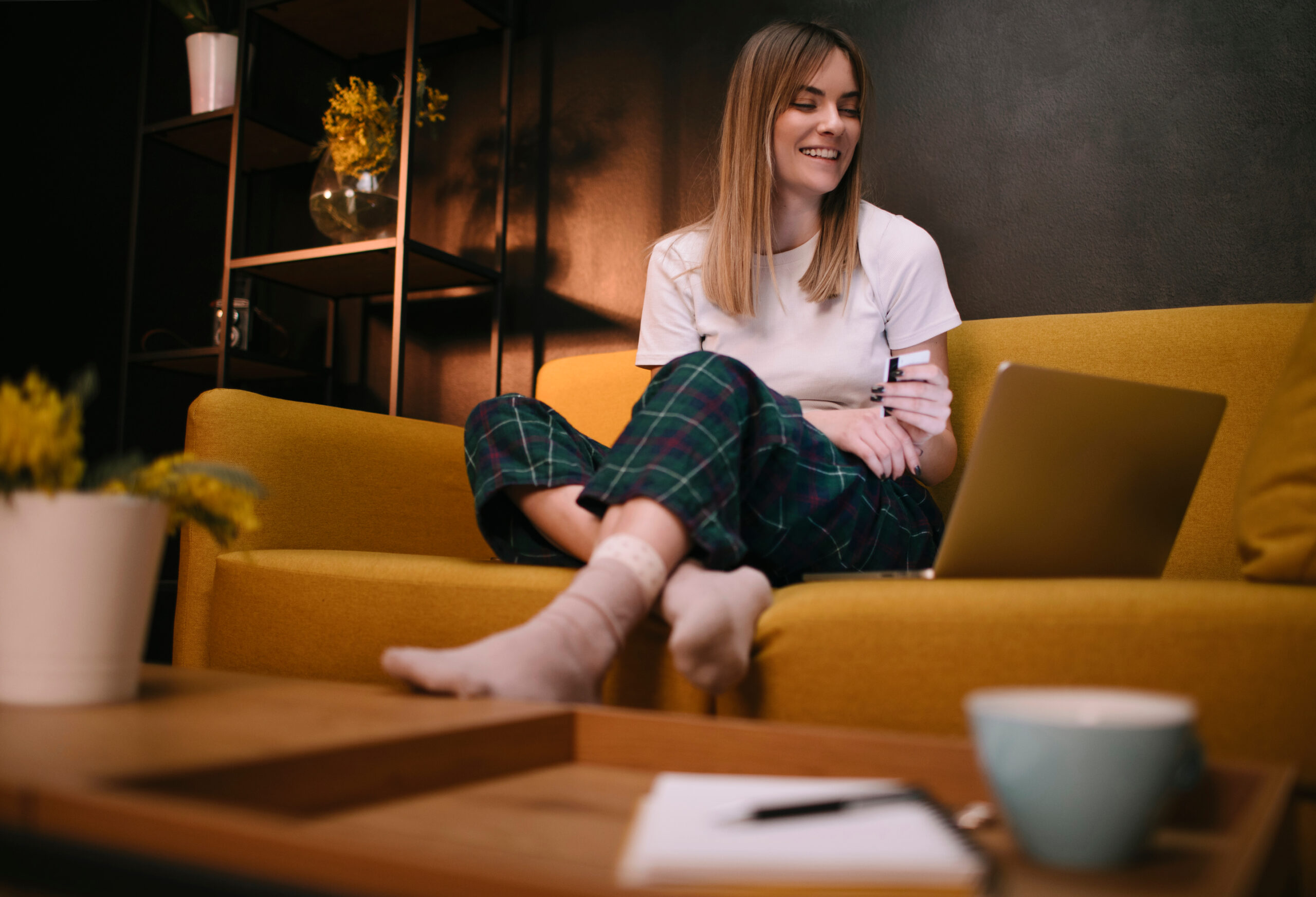 A woman in pajamas sits barefoot on a yellow sofa, smiling at a laptop on the table in front of her. The setting is cozy, with warm lighting and decorative plants in the background.
