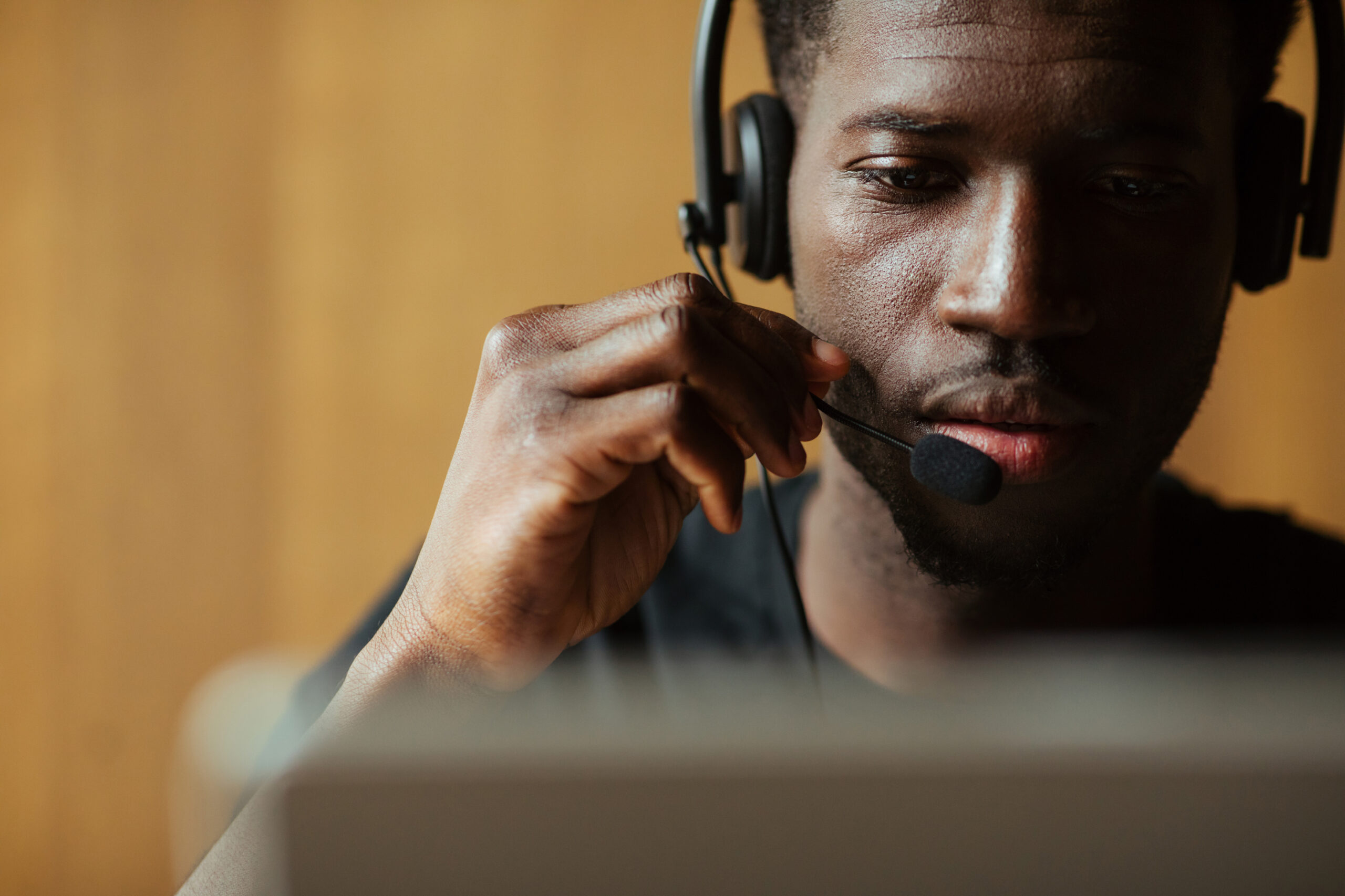 A man wearing a headset with a microphone looks at a computer screen, appearing focused. The background is softly blurred, drawing attention to his face and the headset.