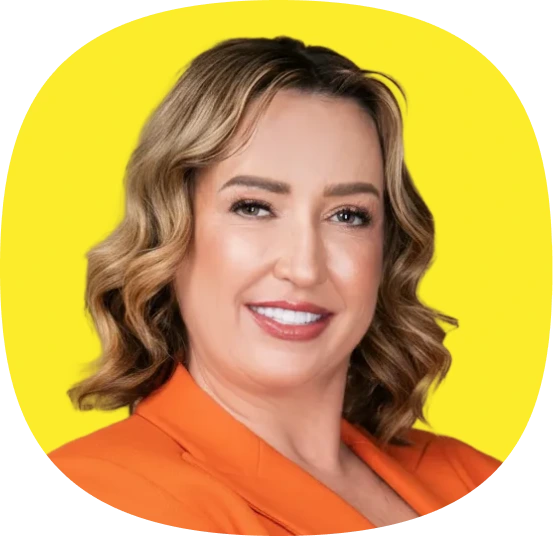 A woman with wavy, shoulder-length blonde hair smiles at the camera. She is wearing an orange blazer and is posed in front of a bright yellow background.