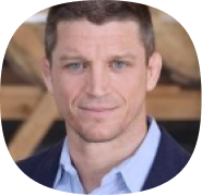 A man with short brown hair and light eyes, wearing a blue collared shirt and dark blazer, is looking at the camera and smiling slightly. The background is blurred with wooden elements visible.