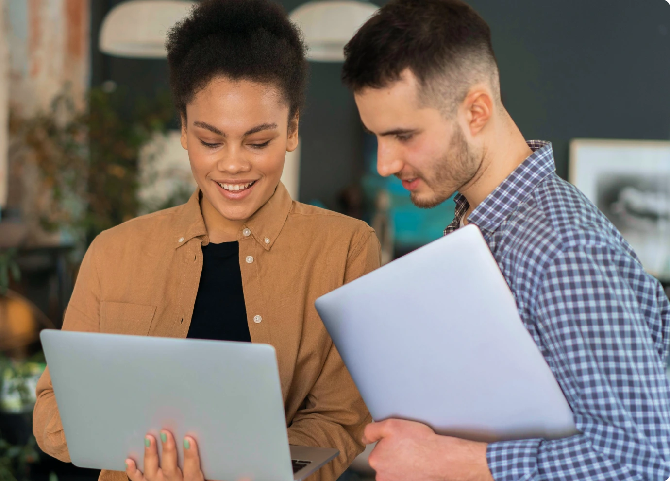 Two people standing together indoors, smiling and looking at a laptop screen. One person holds the laptop while the other holds a closed laptop in their arm. Both appear engaged and focused on the device.