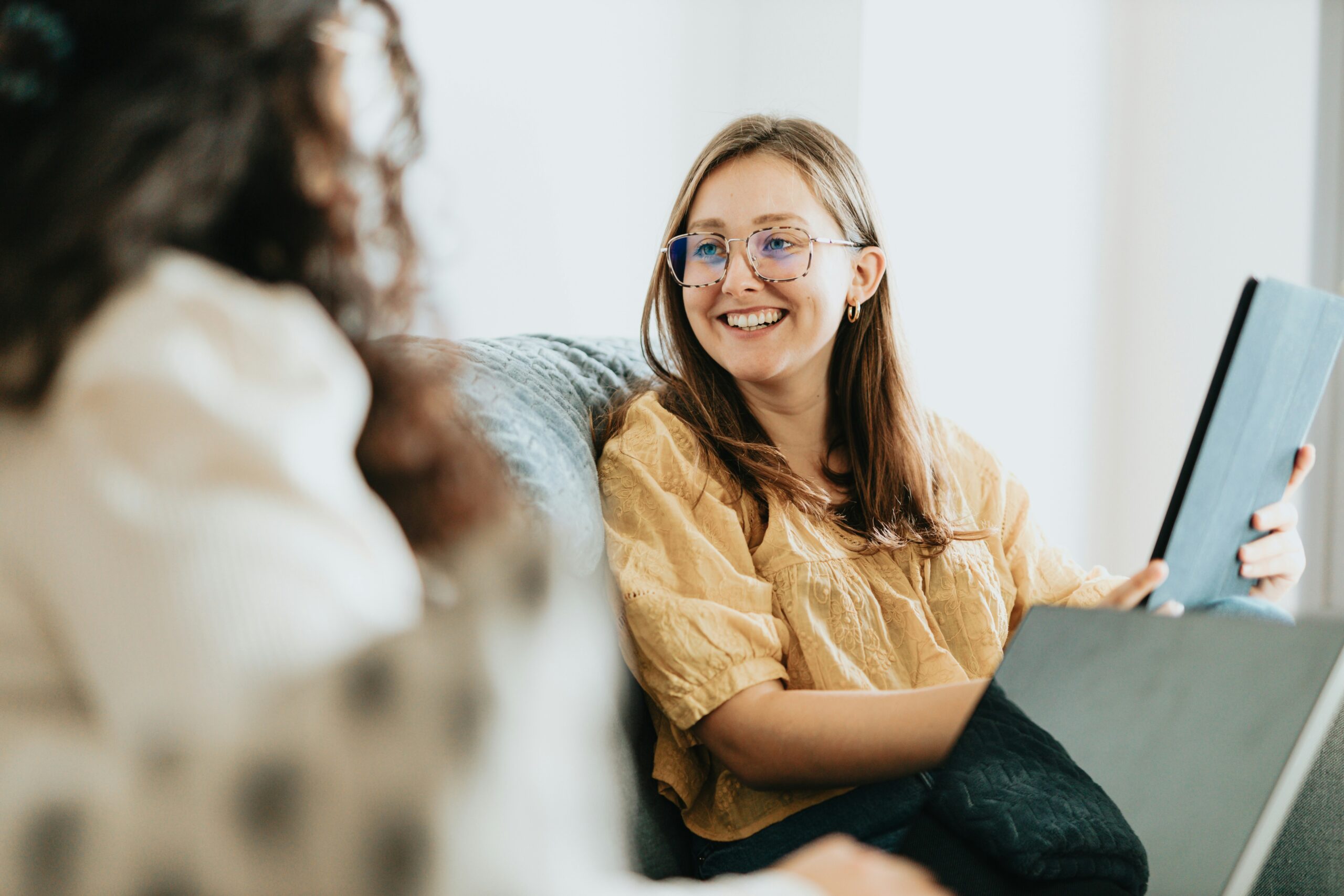 Two people sit on a couch having a conversation. One, smiling in glasses and a yellow top, holds a tablet—perhaps reviewing zoho zia alternatives. The other person is partially visible, facing her and listening attentively.