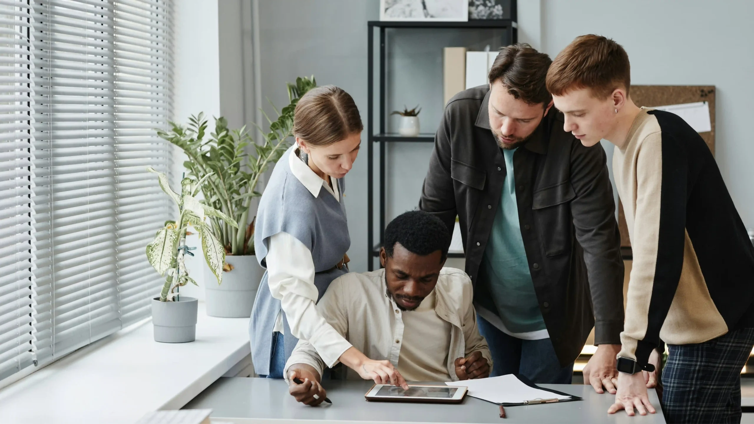 Four people gather around a desk, looking at a tablet. One person is seated and touching the tablet while the others stand close, observing proactive customer service strategies. Papers, a notebook, and plants are visible on the desk and nearby shelves.