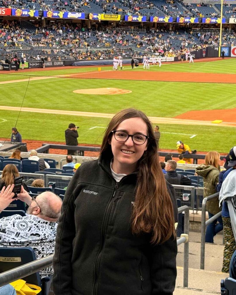 A woman with long brown hair and glasses smiles at the camera in a stadium seat at a baseball game, capturing authentic fan experiences with the field, players, and an enthusiastic crowd in the background.